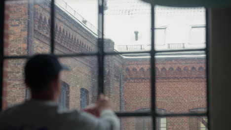 Person-gazes-thoughtfully-through-a-snow-dappled-window,-historic-brick-building-backdrop
