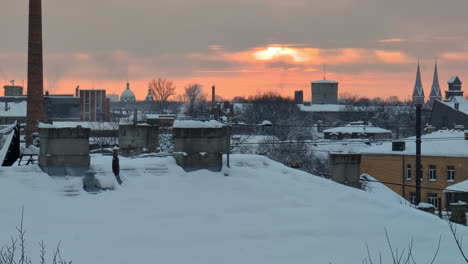 Schnee-Fällt-In-Zeitlupe-Auf-Das-Dach-Des-Städtischen-Industriegebiets,-Während-Die-Sonne-Am-Grauen-Winterhimmel-Orange-Leuchtet