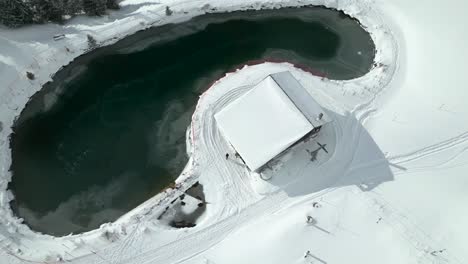 Aerial-drone-zoom-in-shot-over-a-partly-frozen-lake-beside-a-resort-building-in-Engelberg-Brunni-bahnen-in-Switzerland-on-a-sunny-day