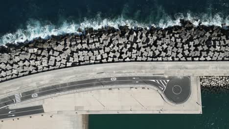 Ocean-waves-crash-on-large-breakwater-jetty-protecting-marine-port-and-road,-drone-top-down
