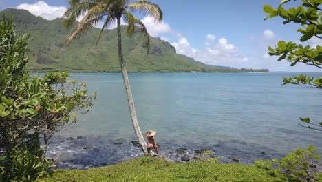 Una-Mujer-En-Bikini-Rosa-Y-Sombrero-De-Paja-Posa-Junto-A-Una-Palmera-En-Una-Playa-Hawaiana,-Con-Un-Hermoso-Mar-De-Fondo