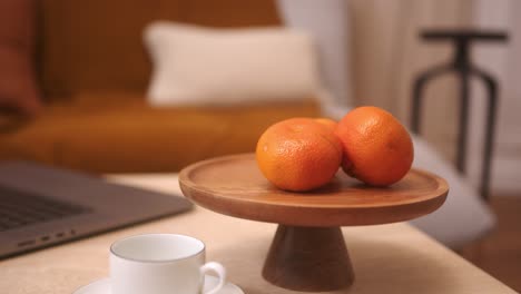 Two-Oranges-on-Wooden-Cake-Stand-Beside-White-Mug-and-Laptop