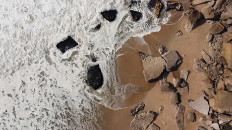 Luftaufnahme-Aus-Der-Vogelperspektive-Auf-Meereswellen,-Die-An-Einem-Felsigen-Und-Steinigen-Strand-In-Nazare,-Portugal,-Zusammenbrechen