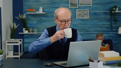 Tired-elderly-man-sitting-at-desk-drinking-coffee,-reading-and-typing-at-laptop