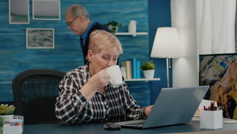 Older-mature-business-woman-using-laptop-sitting-at-workplace-drinking-coffee