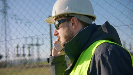 Male-engineer-listening-over-the-radio-while-nodding-his-head-at-the-electric-substation,-handheld-closeup