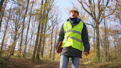 male-engineer-in-safety-vest-and-hard-hat-walking-in-the-woods-alone-while-carrying-clipboard,-handheld-tracking-shot