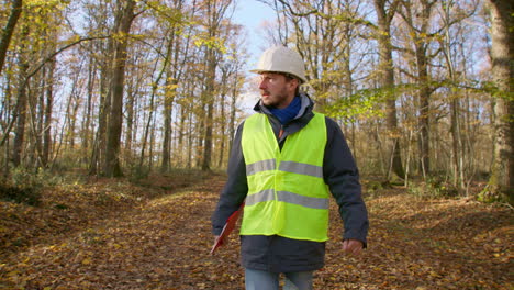 Male-engineer-walking-alone-towards-the-camera-in-the-forest-while-carrying-clipboard-to-survey-for-trees,-handheld-following