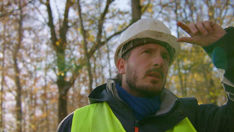 Male-engineer-wearing-hard-hat-staring-upwards-at-the-trees-in-the-middle-of-the-forest,-handheld-closeup