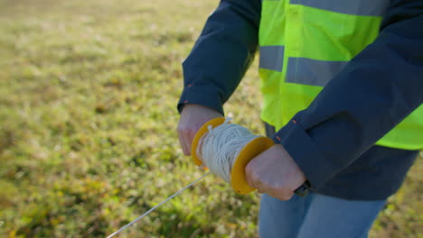 Male-engineer-deploying-a-wire-across-the-grassy-field-from-handheld-spool-of-cable-on-a-rod,-handheld-closeup