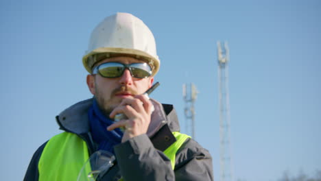 Male-engineer-talking-over-the-radio-with-cellular-towers-in-the-background-while-switching-subject-focus,-closeup