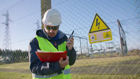 Male-engineer-talking-over-the-walkie-talkie-and-reading-on-clipboard-at-the-electric-substation,-handheld-dynamic