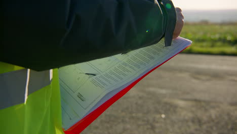 Engineer-wearing-safety-reflective-vest-reading-the-manual-on-the-clipboard-while-holding-a-pen,-handheld-closeup