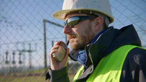Male-engineer-listening-over-the-walkie-talkie-while-nodding-his-head-at-the-electric-substation,-handheld-closeup