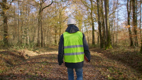 Male-engineer-walking-alone-in-the-forest-while-carrying-clipboard-in-safety-vest-to-survey-for-trees,-handheld-following