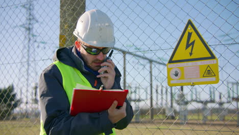 Male-engineer-talking-over-the-radio-while-holding-a-clipboard-at-the-electric-substation,-handheld-dynamic