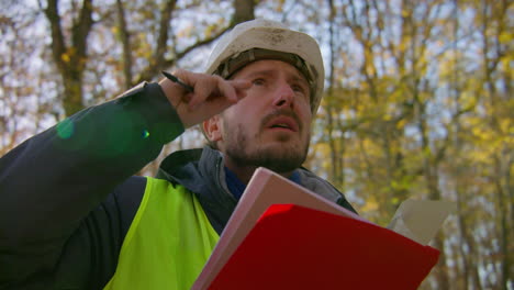 Male-engineer-looking-upwards-at-the-trees-and-reading-on-the-clipboard-in-the-middle-of-the-forest,-handheld-closeup