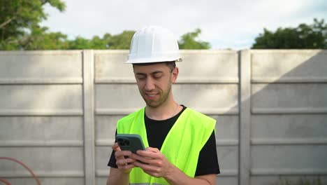 A-Manager-Wearing-a-White-Hard-Hat-is-Using-His-Phone-for-Texting-While-Overseeing-the-Progress-of-the-Work---Medium-Close-Up