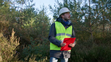 Male-engineer-looking-around-the-trees-at-the-forest-while-holding-on-to-his-clipboard,-handheld