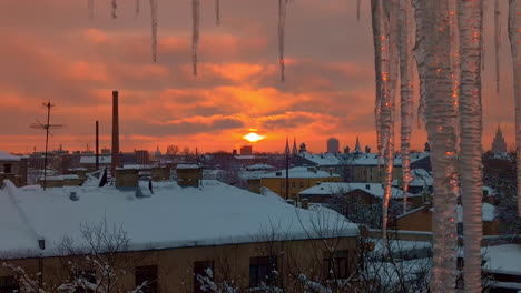 Roter-Sonnenuntergangshimmel-Und-Verschneite-Stadtdächer-Mit-Eiszapfen-Im-Vordergrund