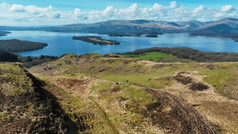 Views-of-Scotland,-Cinematic-Aerial-of-Couple-Overlooking-Loch-Lomond-on-Conic-Hill-on-Sunny-Spring-Day