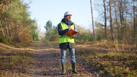 male-engineer-opening-his-notes-on-his-clipboard-and-writing-on-it-while-in-the-middle-of-the-forest,-handheld