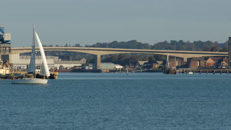 Kleines-Segelboot,-Das-Von-Motorisierten-Passagierkatamaranen-In-Solent,-Southampton,-Mit-Der-Itchen-Mautbrücke-Im-Hintergrund-überholt-Wird