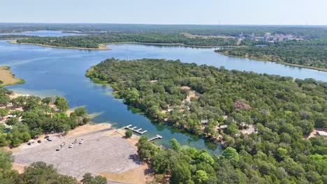 Moving-above-a-boat-ramp-parking-lot,-to-the-left-above-a-pretty-blue-lake-and-recreational-area-that-has-shoreline-and-a-campsite-on-the-other-side