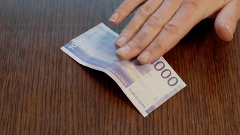 Hand-Placing-Norwegian-1000-kr-Note-in-center-of-Wooden-Table