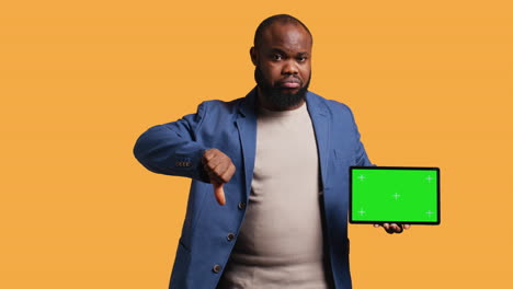 Man-showing-thumbs-down-sign-gesturing-holding-green-screen-tablet