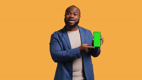 Smiling-influencer-presenting-green-screen-phone,-studio-background