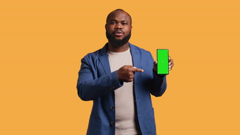 Man-presents-phone-with-green-screen-display,-isolated-over-studio-background