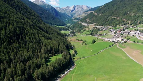Green-valley-between-huge-mountain-ranges-with-fields-and-a-small-village-under-a-blue-sky,-drone-panoramic-view