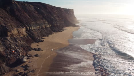 Cliffside-beach-waves-in-Portugal,-morning-glow---Aerial