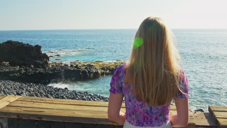 Blonde-girl-standing-and-looking-over-calm-sea-on-summer-day,-rear-view