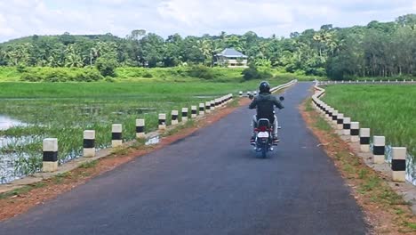 Usando-Zapatos-Con-La-Marca-Z,-Un-Hombre-Usa-Un-Casco-En-Una-Motocicleta-Y-Pasa-Por-Un-Camino-Rural-En-La-India,-Viajando-En-Una-Vieja-Motocicleta