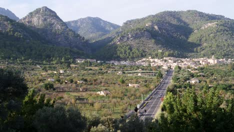 Traveling-cars-on-a-serene-straight-road-to-picturesque-Caimari-village,-embraced-by-lush-greenery-and-rolling-hills-on-a-beautiful-autumn-evening-in-Mallorca