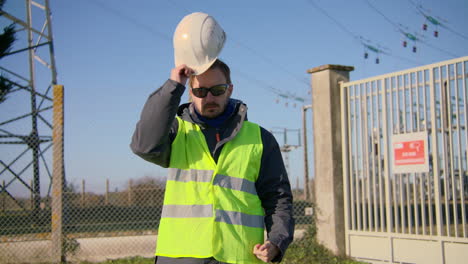 Male-engineer-closing-his-jacket-zipper,-wears-hard-hat,-and-crossing-his-arms-at-the-electric-substation,-handheld