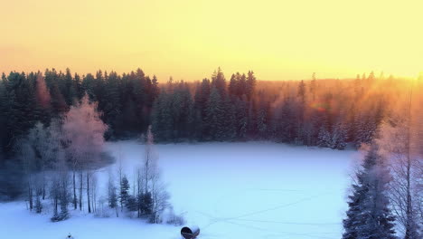 Remote-sauna-covered-in-snow-and-bright-sunset-above-forest,-aerial-view