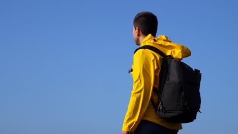 Nachdenklicher-Backpacker-Mann-Mit-Gelbem-Regenmantel,-Der-An-Windigen-Tagen-In-Den-Blauen-Himmel-Blickt