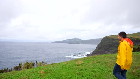 Man-in-yellow-raincoat-exploring-grassy-volcanic-cliffs-by-the-Atlantic-at-Castelo-Branco,-Faial