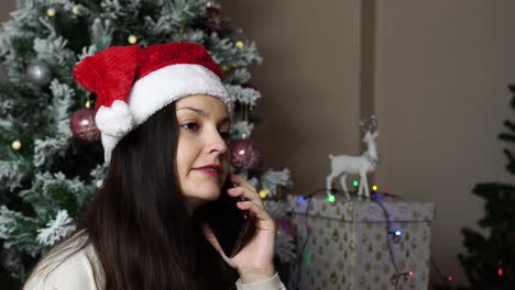 Schöne-Brünette-Mit-Weihnachtsmütze-Telefoniert-In-Der-Nähe-Von-Weihnachtsbaum-Und-Geschenken