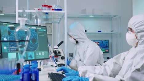 Doctor-in-ppe-suit-working-at-pc-while-chemist-using-microscope