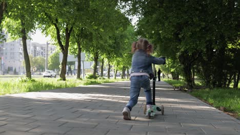 Little-girl-rides-scooter-in-green-pring-park-with-mother-and-grandparents