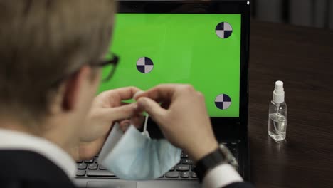 Mann-Nimmt-Medizinische-Maske-Ab,-Nimmt-Desinfektionsmittel-Und-Benutzt-Es-In-Der-Nähe-Eines-Laptops-Mit-Grünem-Bildschirm