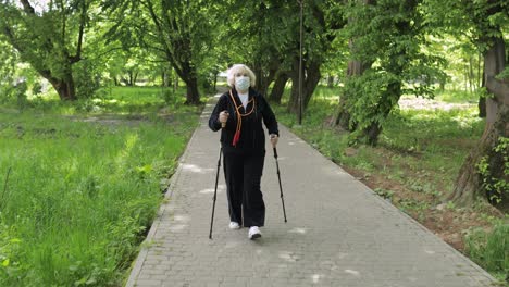 Active-senior-old-woman-in-mask-training-Nordic-walking-in-park-at-quarantine