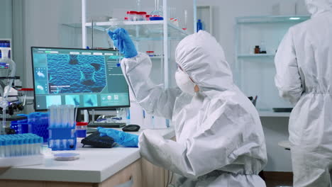 Scientist-nurse-in-ppe-suit-taking-notes-on-computer-looking-at-test-tube