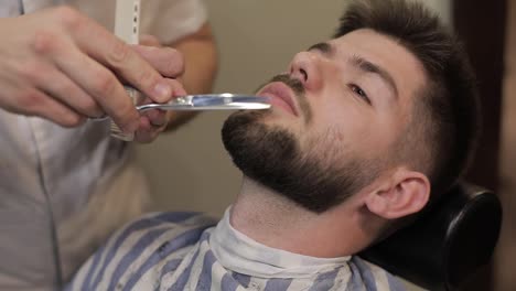 Client-with-black-beard-during-beard-shaving-in-barber-shop.-Groom,-masculine