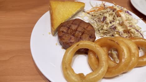 Close-up-of-a-plate-filled-with-onions-rings,-toasted-bread,-coleslaw-salad,-and-a-cut-of-medium-rare-burger-steak,-served-in-a-restaurant-in-Bangkok,-Thailand