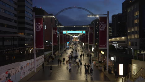 At-dusk-people-walking-along-Orient-Way,-that-links-Wembley-Park-Underground-train-station-to-Wembley-Stadium,-pass-new-build-apartment-blocks-and-large-signs-on-poles-advertising-rental-properties